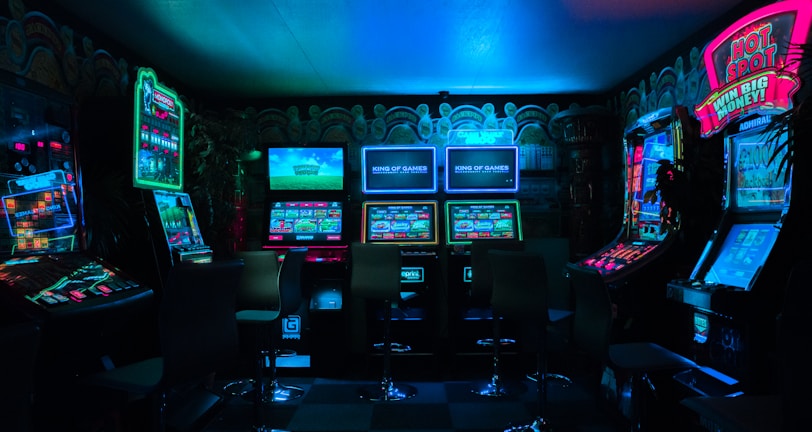 Dimly lit gaming arcade featuring a variety of brightly illuminated slot machines and video gaming screens. Neon signs and colorful displays create a vibrant atmosphere. The arrangement of gaming machines suggests an inviting space for entertainment, with chairs positioned for players.