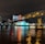 A brightly lit casino with neon signs and red lights is reflected in a river. A bridge stretches across the water, with an urban skyline visible in the background against an overcast night sky.