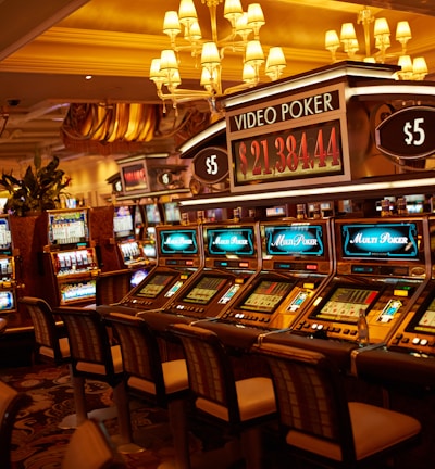 A casino floor featuring rows of brightly lit video poker machines. The area is illuminated by elegant chandeliers, with a display board showing a large sum of money, suggesting a possible jackpot. The rich decor includes luxurious carpets and comfortable seating, adding to the opulent atmosphere.