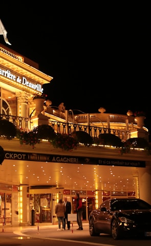 A luxurious casino at night, illuminated with warm, golden lights. The building features elegant architecture with columns and decorative elements. A few people are walking towards the entrance, and there are a couple of parked cars nearby.