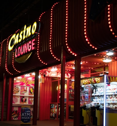 A brightly lit casino lounge entrance adorned with neon lights. The exterior features colorful signage including the words 'Casino' and 'Lounge' in vivid colors, with additional lights framing the building. Inside, a glimpse of a claw machine filled with plush toys and arcade games is visible. The area exudes an inviting yet bustling atmosphere typical of entertainment venues.
