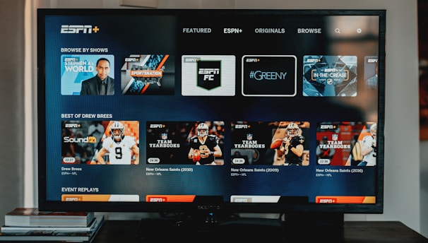 A television screen displaying the ESPN+ streaming interface with various sports-related shows and categories. The screen includes sections such as 'Browse by Shows' and 'Best of Drew Brees' with thumbnails of sports programs featuring athletes in action and analysts.