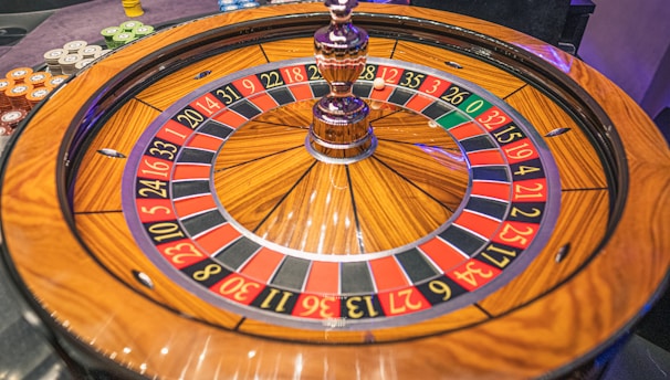 A roulette wheel with alternating red and black numbered pockets on a wooden platform. The wheel contains a small white ball resting in one of the pockets, indicating gameplay. Surrounding the wheel are stacks of colorful poker chips, suggesting a casino environment.