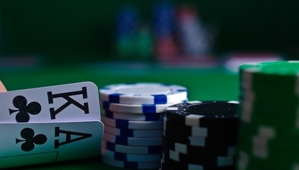 A casino scene featuring two playing cards, the ace and king of clubs, partially concealed by a player's hand. Nearby, there are stacks of multicolored poker chips on a green felt table, with blurred colorful background elements suggesting other chip stacks or cards.