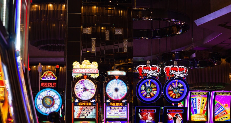A lively casino scene featuring a row of bright, colorful slot machines with themes like Monopoly, Michael Jackson, and Lord of the Rings. The gaming machines display a variety of flashing lights and digital screens, creating an energetic and exciting atmosphere. Reflections of the lights are visible in glossy surfaces above the machines.