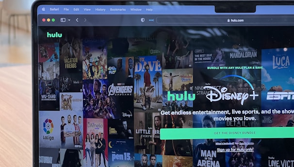 A laptop screen displays a web page featuring a mix of movie and TV show covers, highlighting popular titles and a Hulu and Disney+ bundle promotion. There are visible icons and images of films and shows alongside promotional text offering entertainment and live sports.
