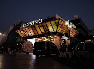 A building with a brightly illuminated entrance is seen at night. The sign above reads 'Casino' in bright letters. The facade features colorful graphics depicting various indulgent themes. Several vehicles are parked near the entrance, and people are visible in the vicinity. Tall lamp posts with spherical lights are present, casting a warm glow.
