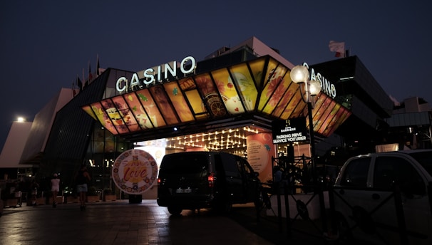 A building with a brightly illuminated entrance is seen at night. The sign above reads 'Casino' in bright letters. The facade features colorful graphics depicting various indulgent themes. Several vehicles are parked near the entrance, and people are visible in the vicinity. Tall lamp posts with spherical lights are present, casting a warm glow.