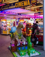 A vibrant amusement arcade featuring various games and bright neon signs. Mechanical horse rides are in the foreground, surrounded by claw machines with plush toys. The entrance is adorned with signs for a cinema and arcade, creating a lively and colorful atmosphere.