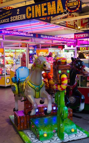 A vibrant amusement arcade featuring various games and bright neon signs. Mechanical horse rides are in the foreground, surrounded by claw machines with plush toys. The entrance is adorned with signs for a cinema and arcade, creating a lively and colorful atmosphere.